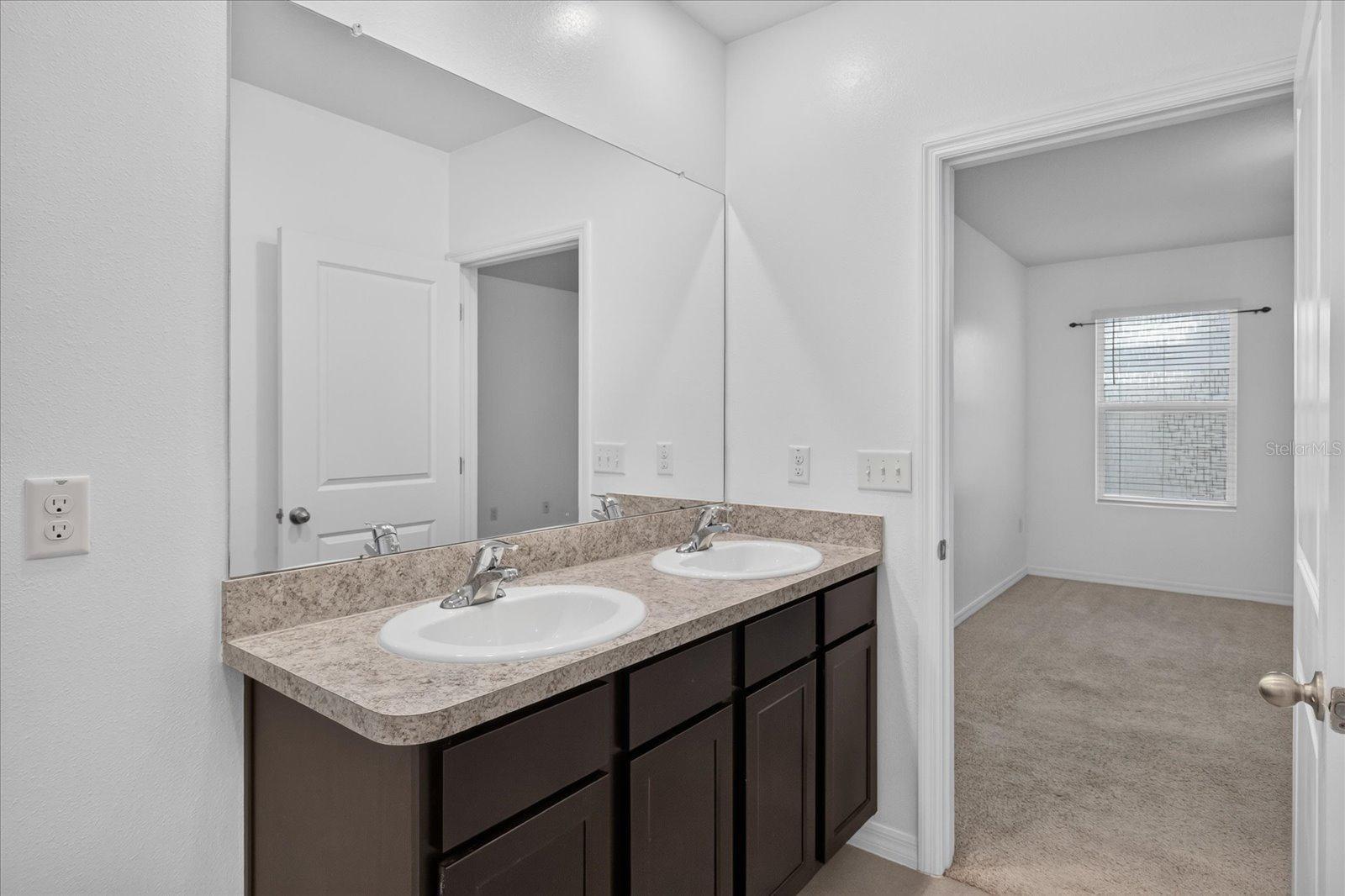Bathroom with a double-sink vanity and large mirror, opening onto an empty carpeted room.