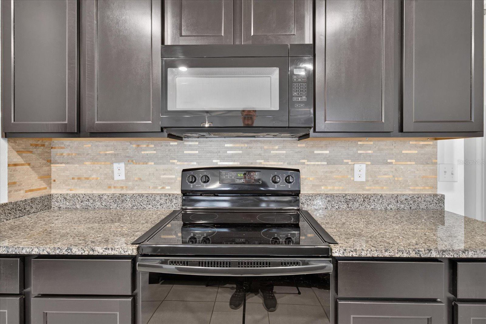 Kitchen with dark cabinets, granite counters, patterned tile backsplash, and black electric range beneath a microwave.
