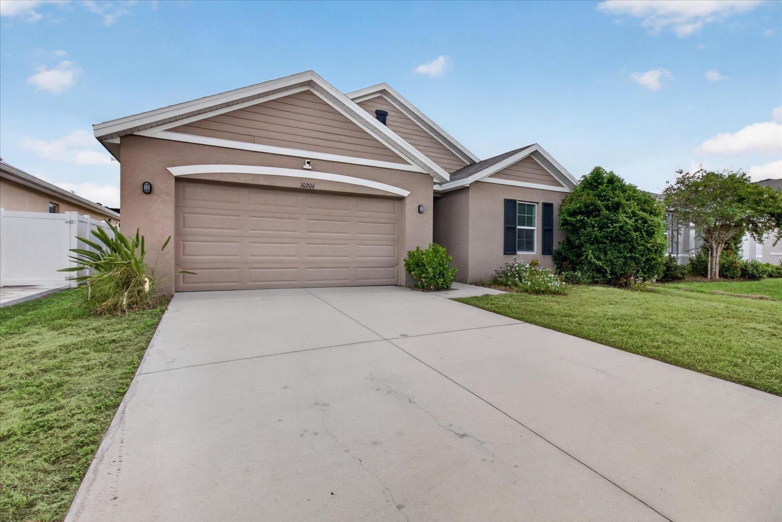 Single-story taupe house with a two-car garage, broad concrete driveway, and white side fence.