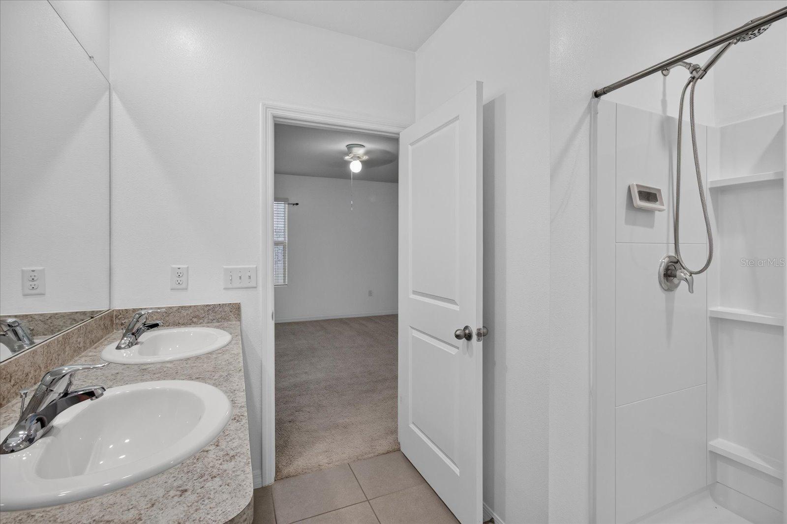 Bathroom with double-sink vanity and tiled shower; open door leads to a carpeted room.