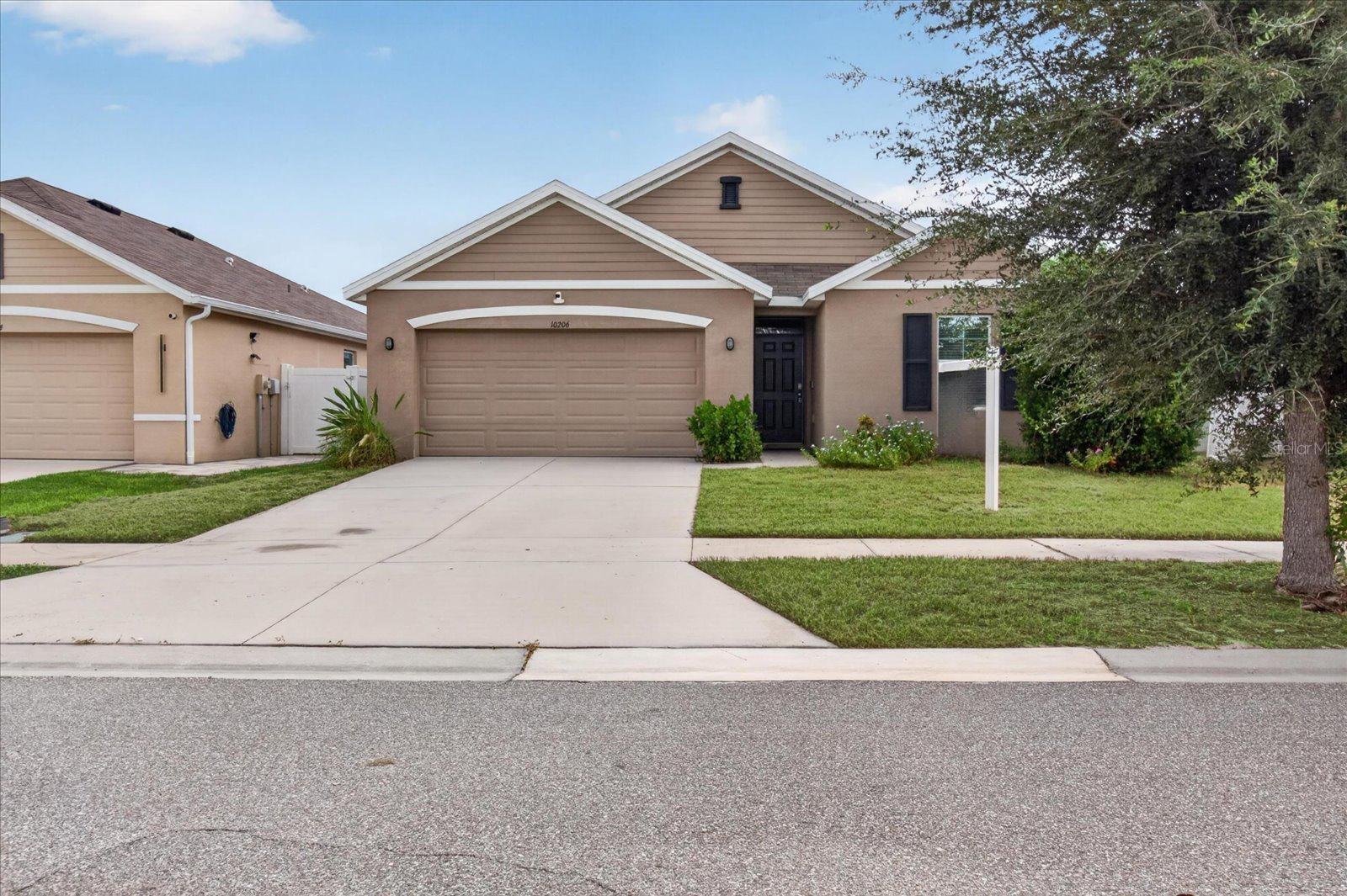 Tan single-story home with a two-car garage, wide driveway, and mature tree in the front yard.