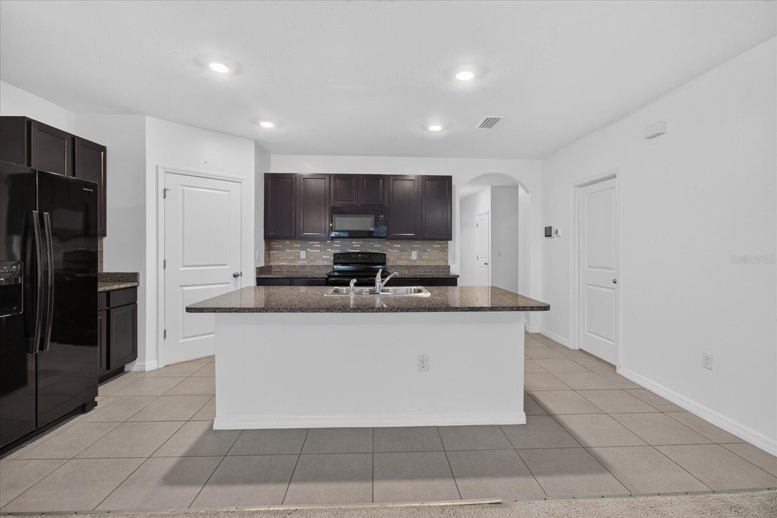 Kitchen with dark cabinets, black appliances, and a large granite island with a sink.