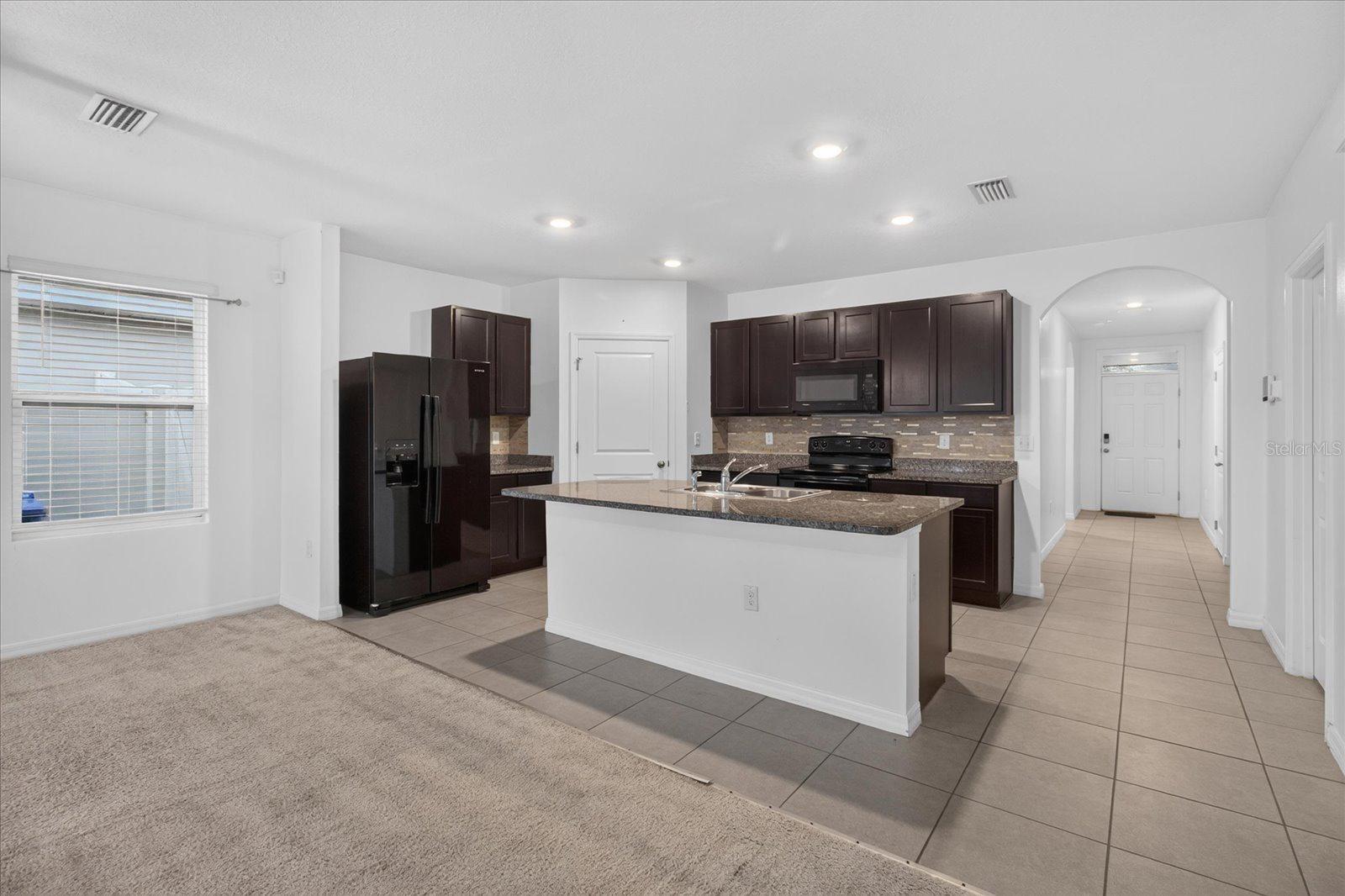 Kitchen with dark cabinets, granite island, stainless steel appliances, and adjoining carpeted living area.