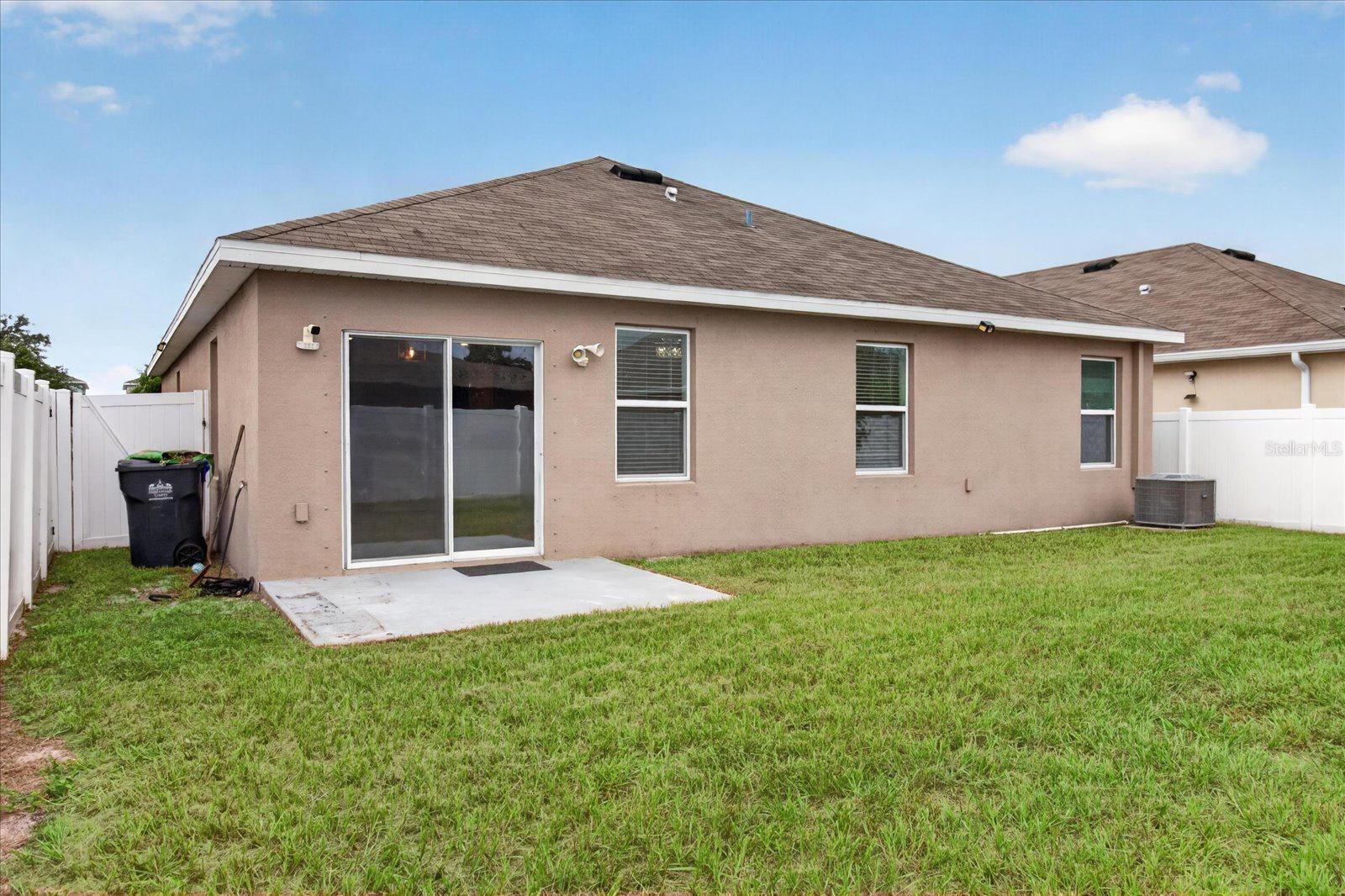 Rear of a stucco home with a sliding glass door, concrete patio, and fenced grassy yard.