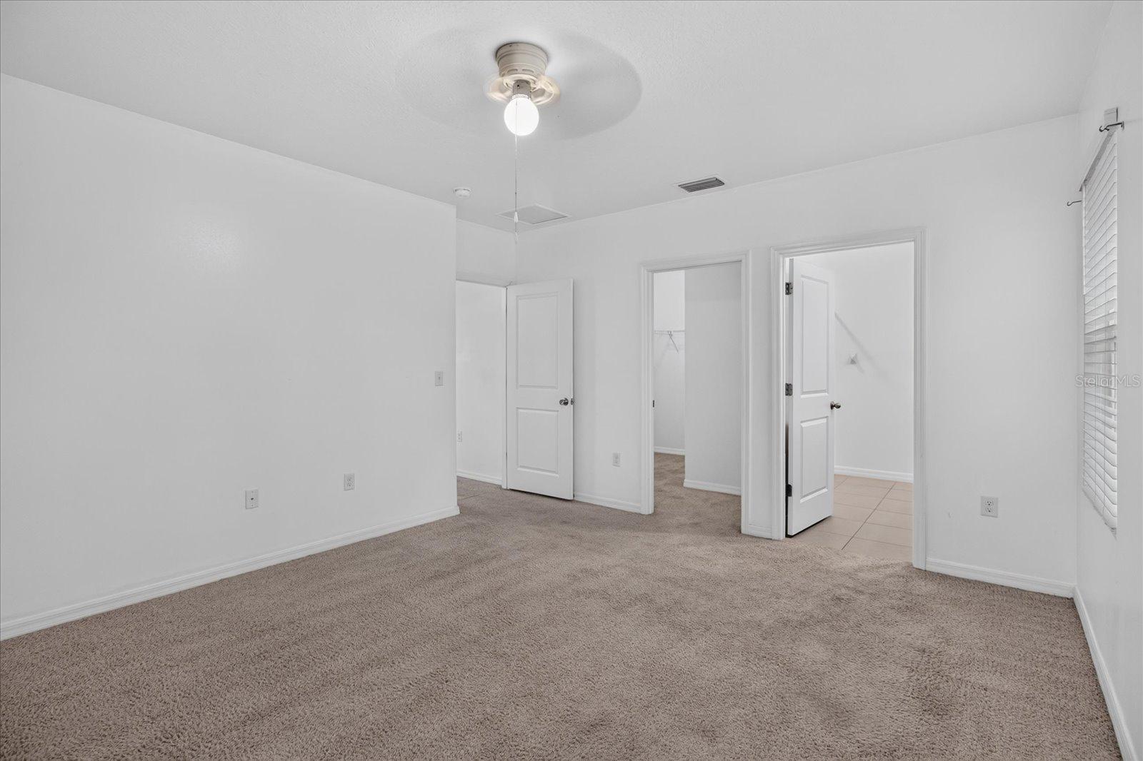 Empty carpeted bedroom with white walls, a ceiling fan, and open doorways.