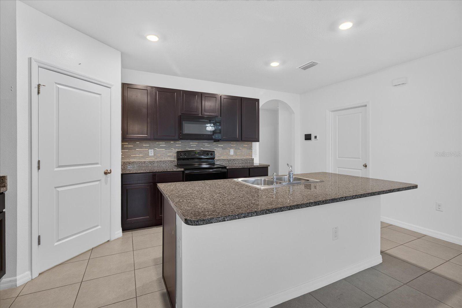 Kitchen with a large granite island, double sink, dark cabinets, and black appliances.