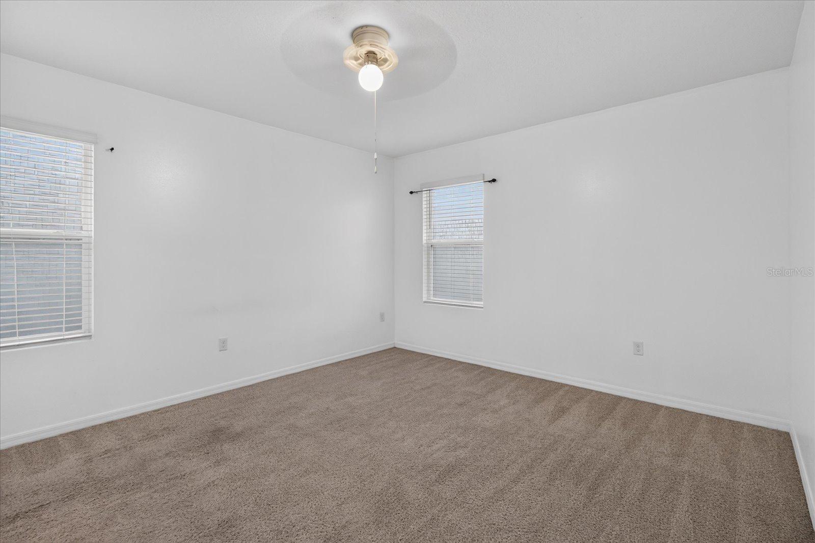Empty bedroom with beige carpet, two blind-covered windows, and a ceiling light.