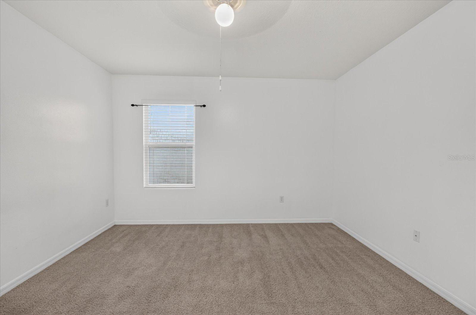 Vacant bedroom with beige carpet, white walls, a window with blinds, and a ceiling fan light.