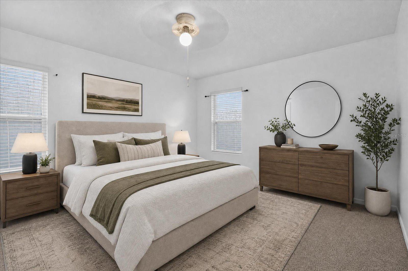 Staged bedroom with an upholstered bed, olive accent bedding, and a wooden dresser beneath a round mirror.