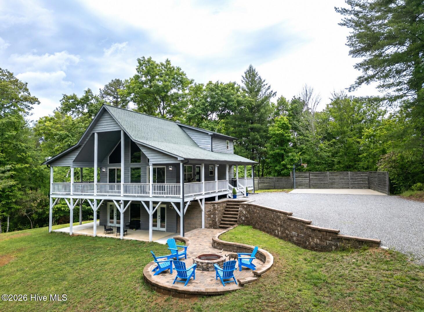 Gray elevated house with a wraparound porch, gravel driveway, and stone fire pit surrounded by blue chairs.