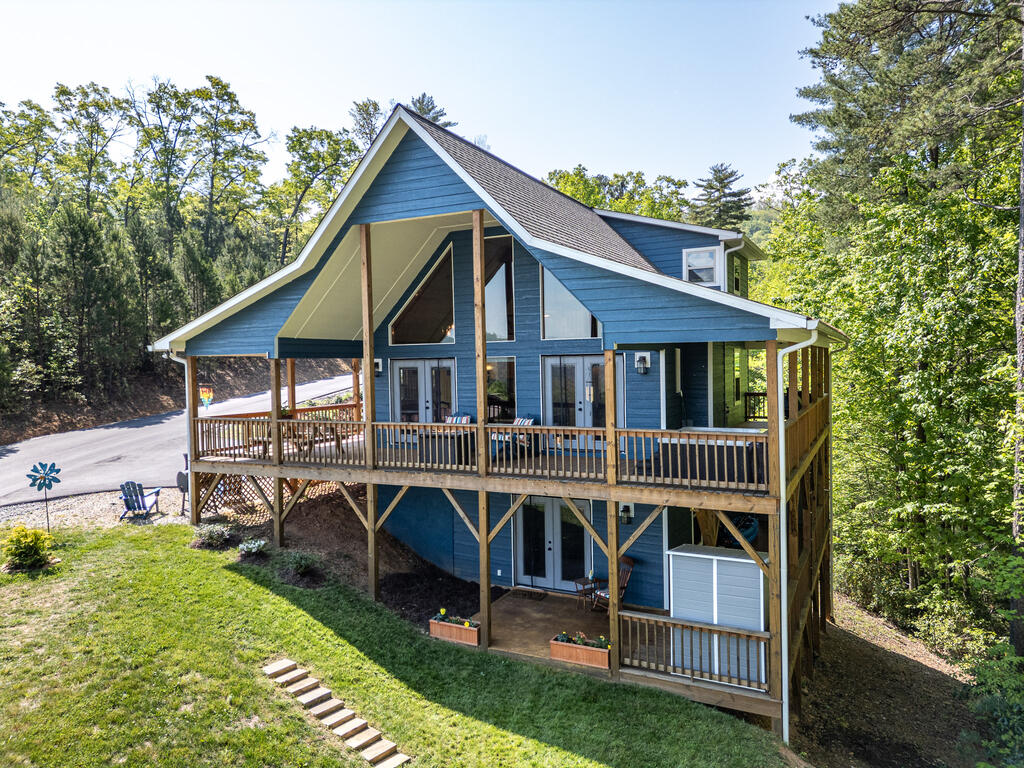 Blue two-story house with a steep gabled roof and elevated wraparound porch among leafy trees.