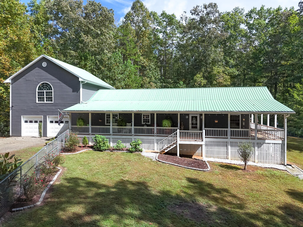 Gray house with a long covered porch, pale green metal roof, and attached garage in a wooded setting.