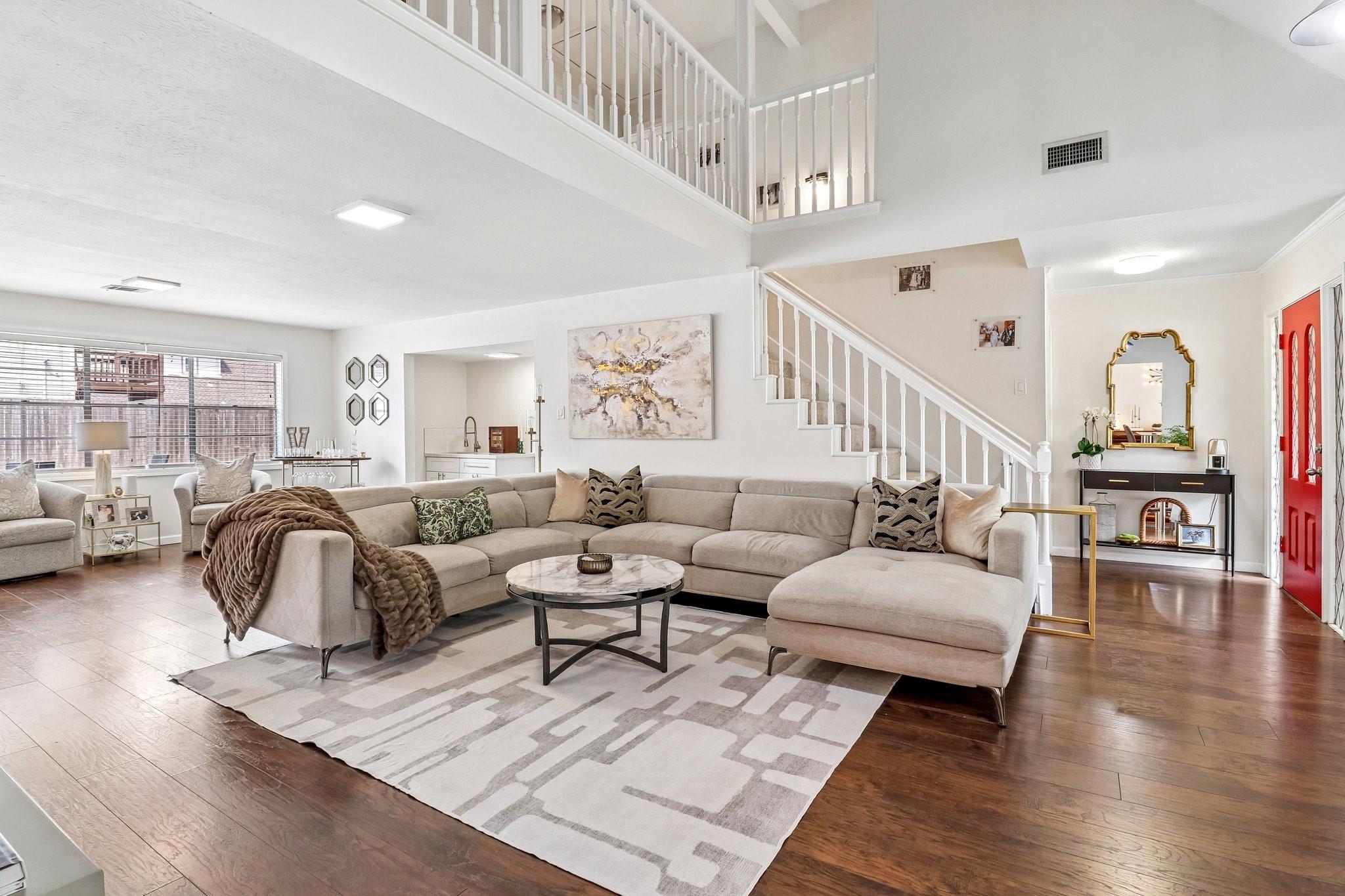 Staged living room with a beige sectional, geometric rug, dark wood floors, and white staircase.