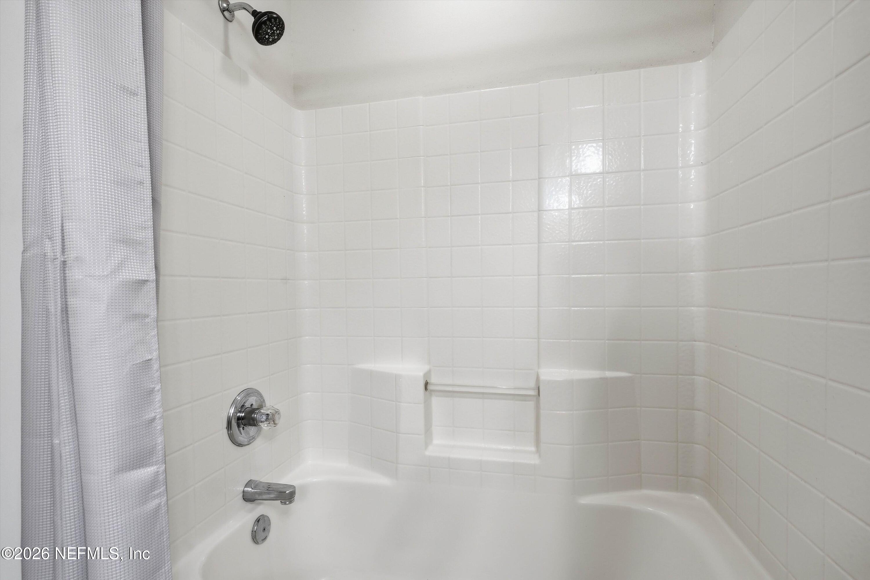 White tiled bathtub and shower with a built-in shelf and chrome fixtures.