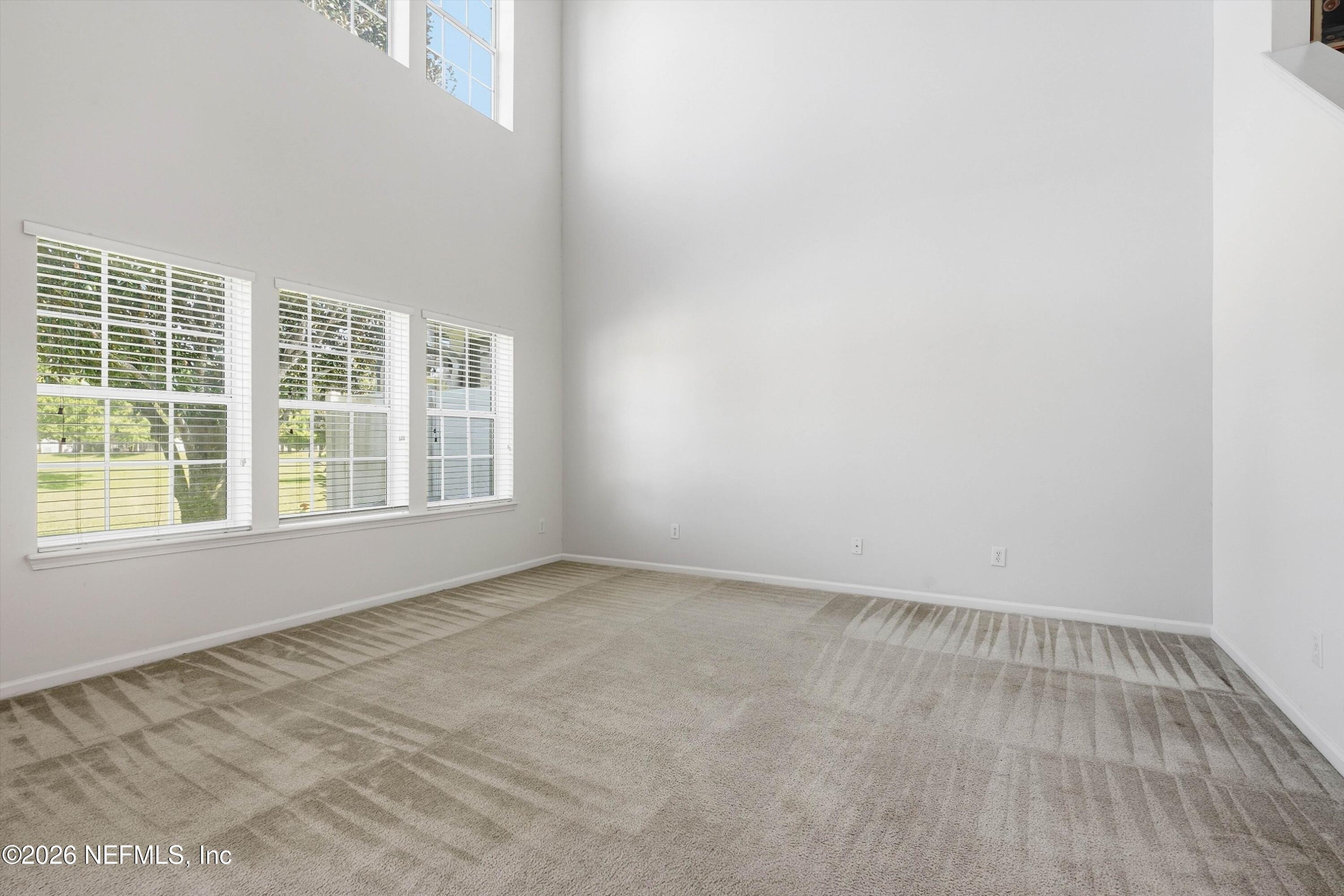Vacant carpeted living room with a high ceiling and large windows casting striped shadows across the floor.