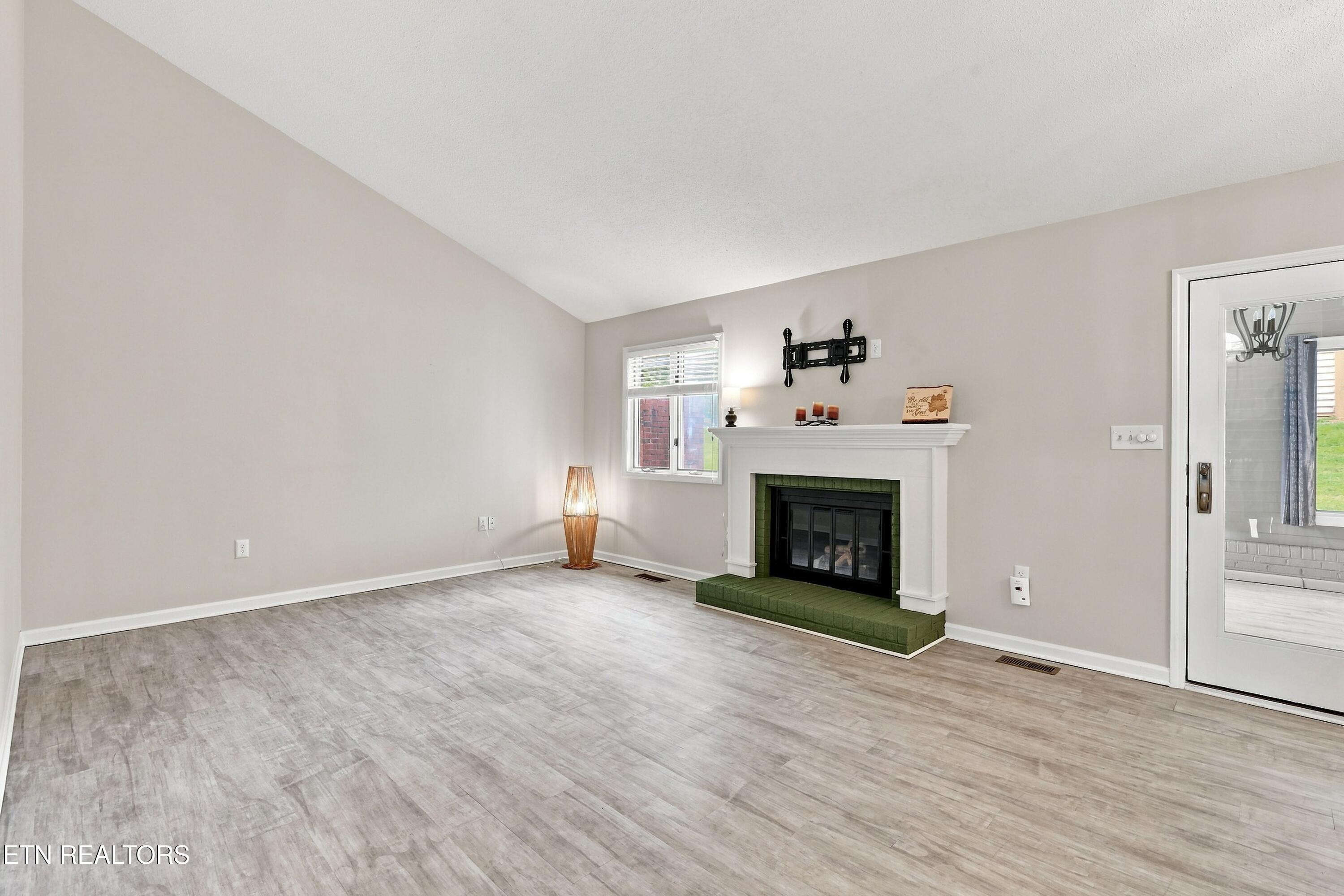 Mostly vacant living room with vaulted ceiling, green-hearth fireplace, and gray plank flooring.