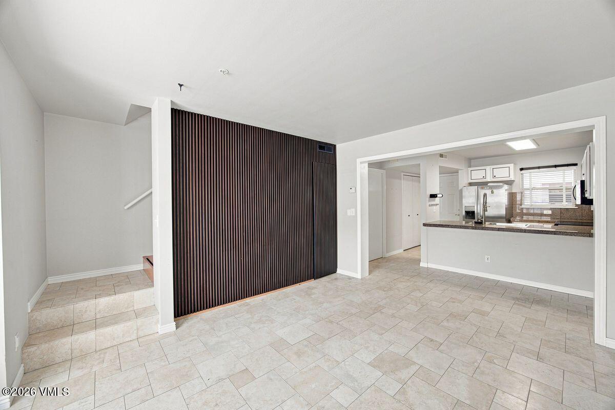 Empty tiled living area with a dark slatted partition and open kitchen.