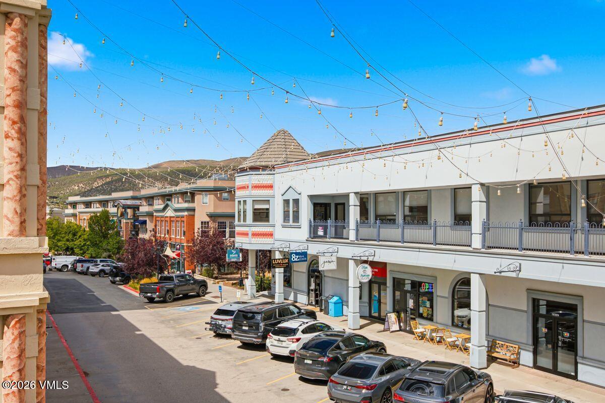 Commercial storefronts with outdoor seating and parked cars beneath string lights, with mountains in the background.