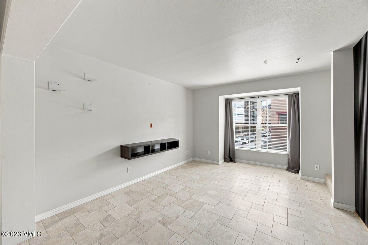 Empty living room with pale tile floors, a floating media shelf, and a large curtained window.