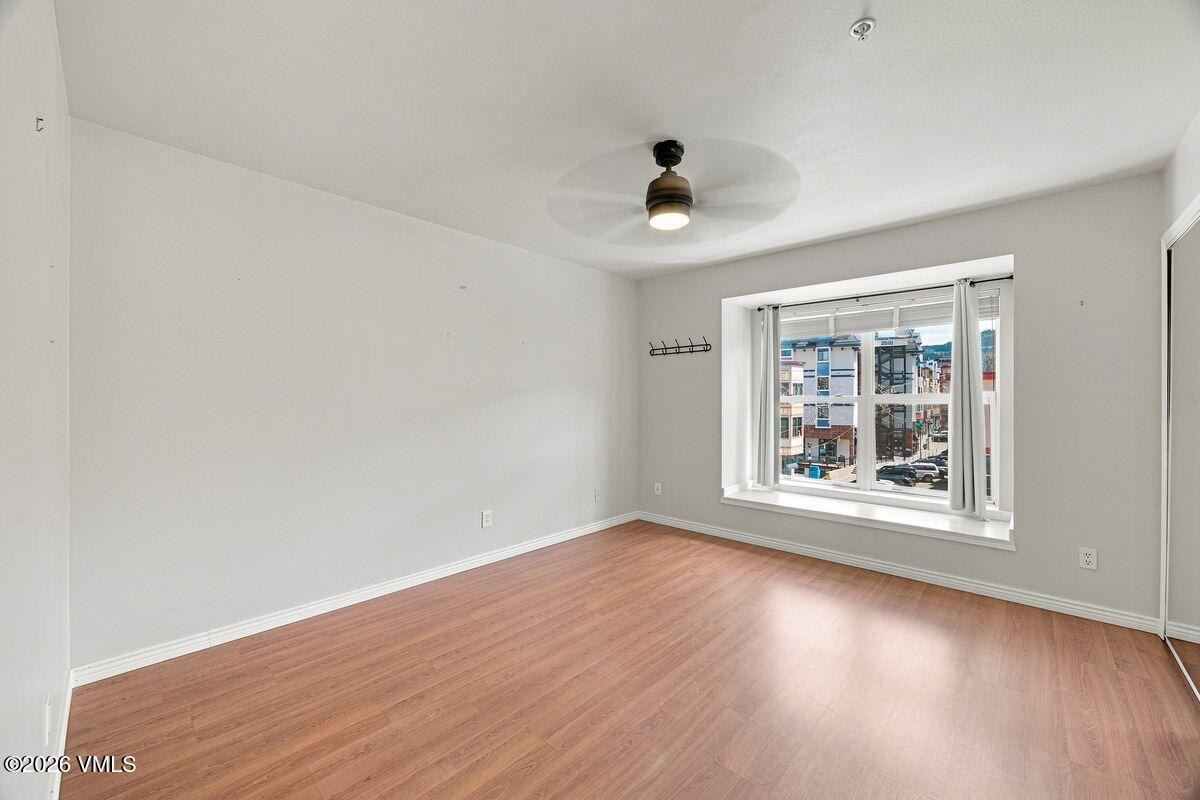 Empty room with wood-look flooring, ceiling fan, and a large window overlooking nearby buildings.