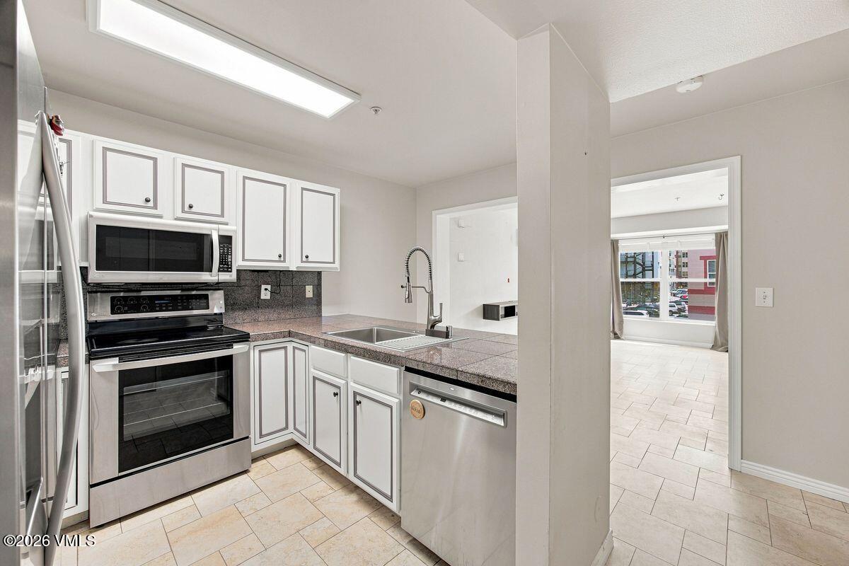 Vacant condo kitchen with white cabinets, stainless steel appliances, and a granite counter opening to a tiled room.