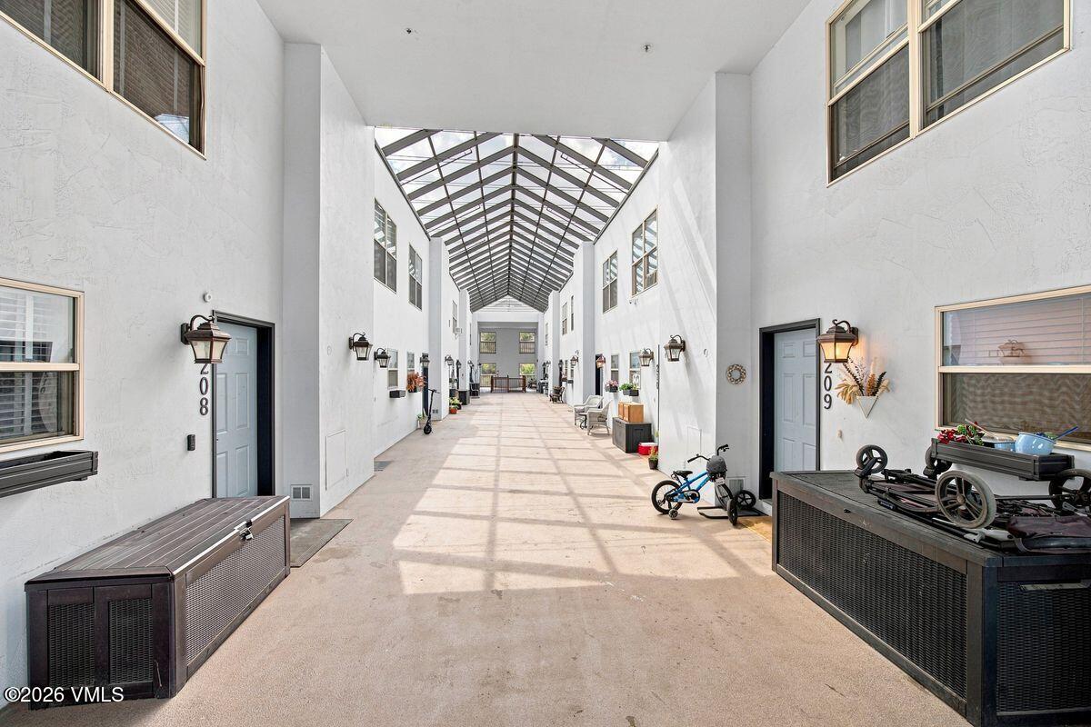 Bright shared corridor beneath a glass-paneled vaulted roof, with unit doors and seating.