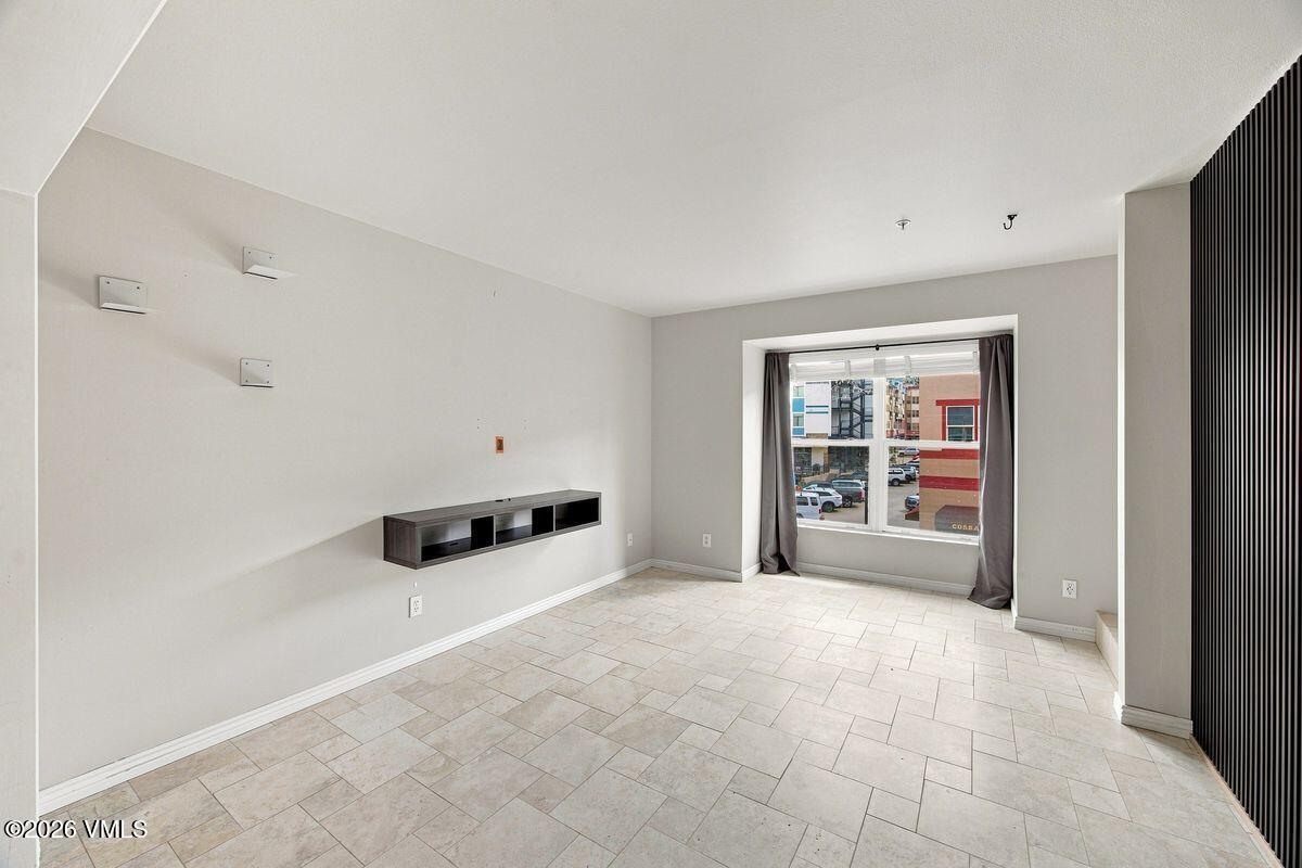 Vacant tiled room with a floating media shelf and a curtained window overlooking nearby buildings.