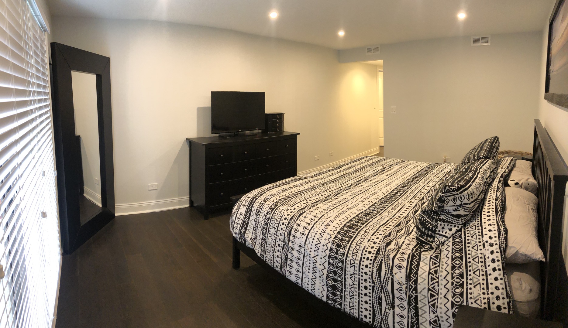 Furnished bedroom with a geometric-patterned bedspread, dark dresser and television, and tall mirror.