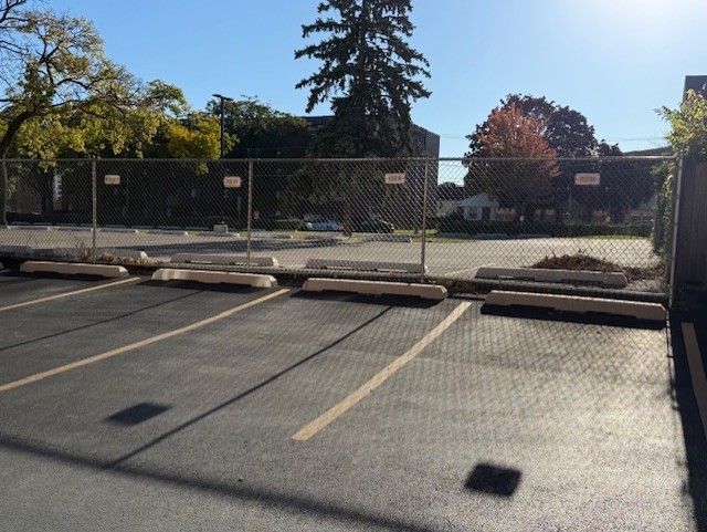 Paved parking lot with marked spaces and concrete wheel stops behind a chain-link fence.