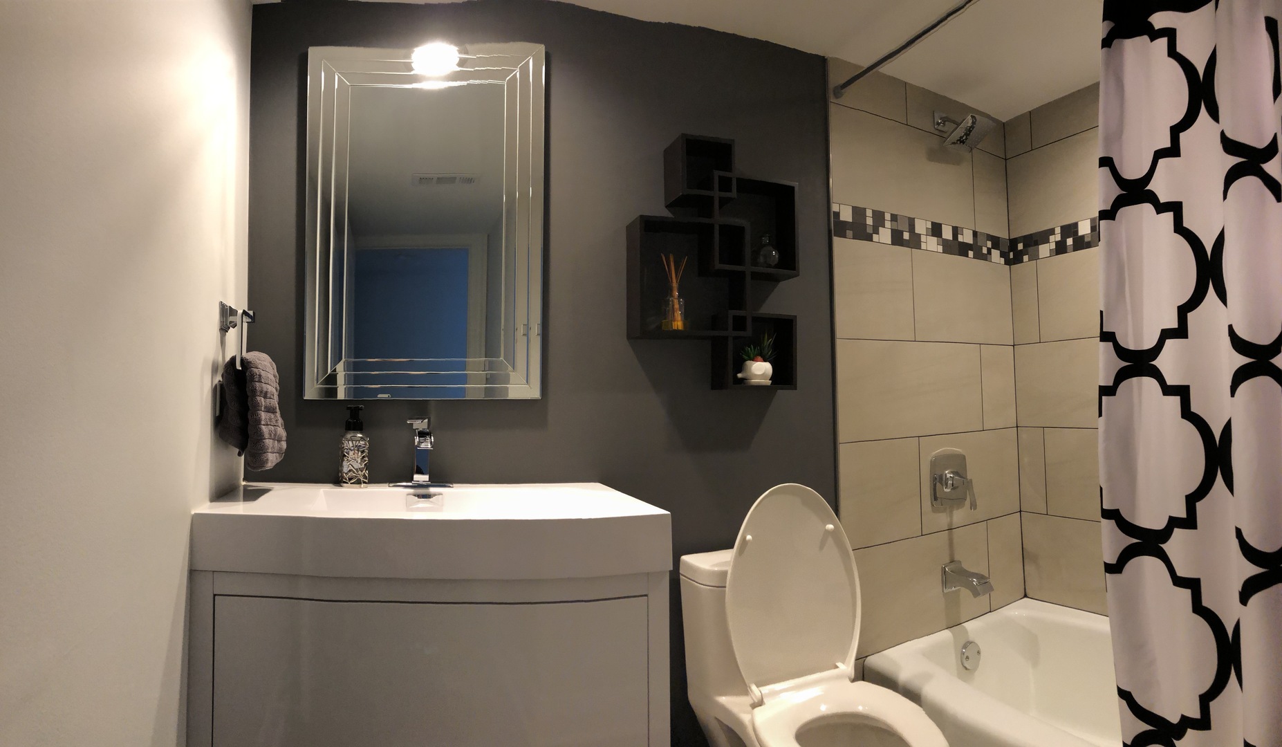 Bathroom with a white vanity, dark accent wall, tiled bathtub, and patterned shower curtain.