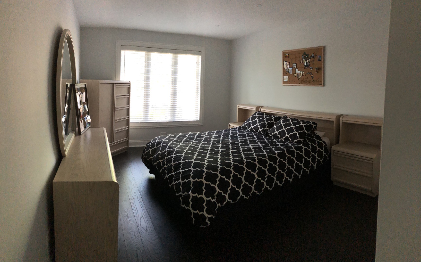 Furnished bedroom with a large patterned bed, light wood furniture, and a bright window with blinds.