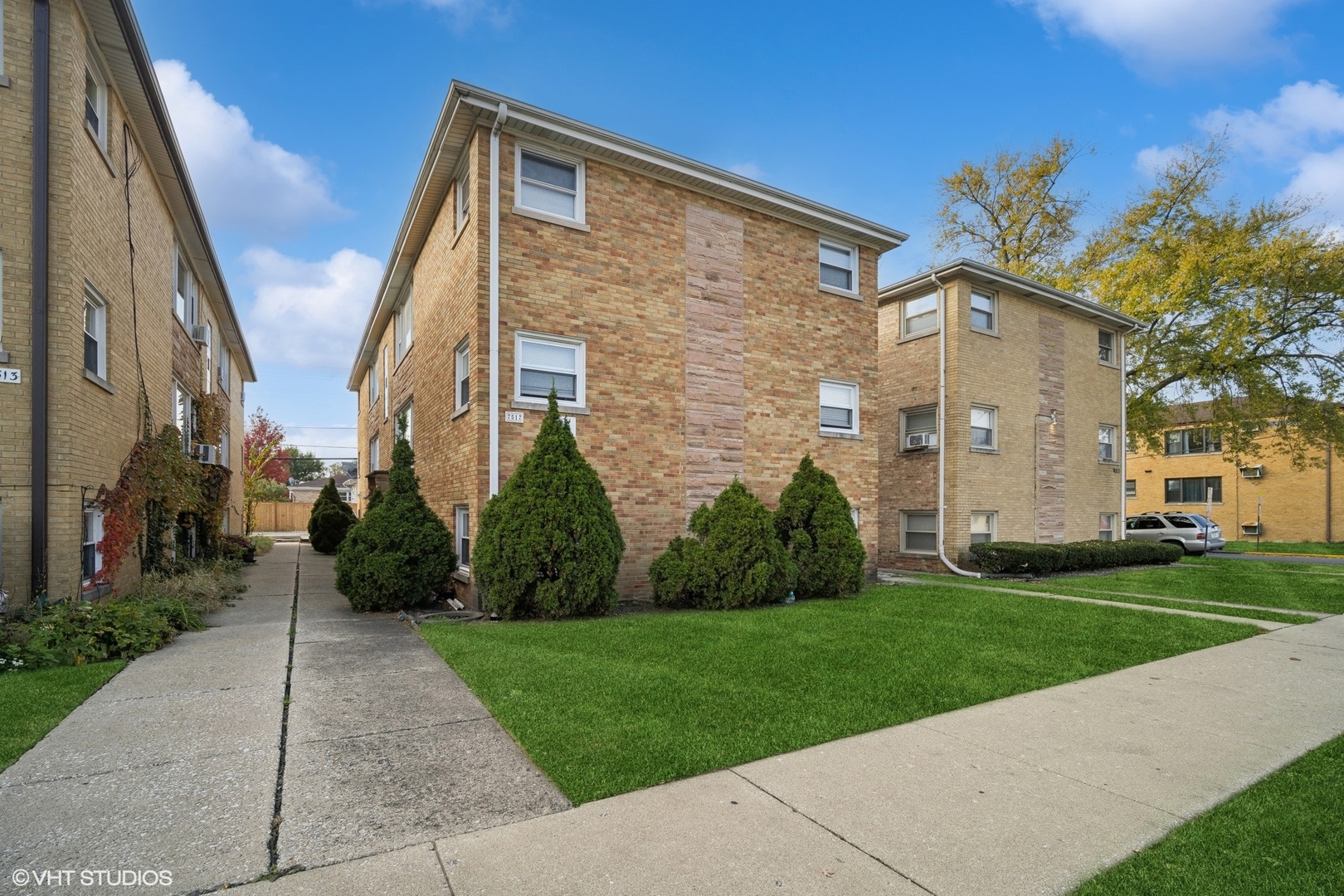 Three-story brick apartment building with front lawn, shrubs, and a concrete walkway.