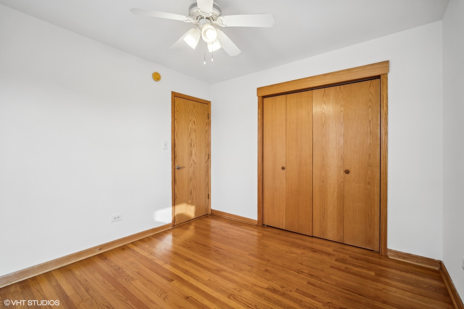 Empty bedroom with hardwood floors, wooden closet doors, and a ceiling fan with light.