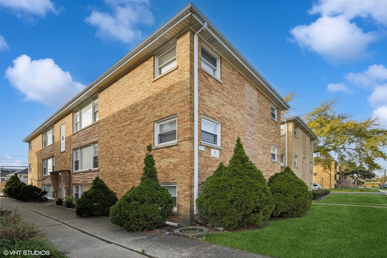 Two-story brick apartment building with trimmed shrubs and a lawn along the corner walkway.