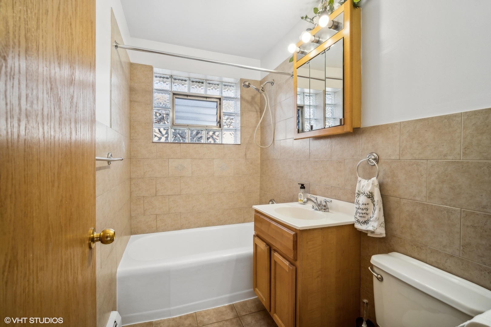 Tiled bathroom with bathtub, glass-block window, wood vanity, mirrored cabinet, and toilet.