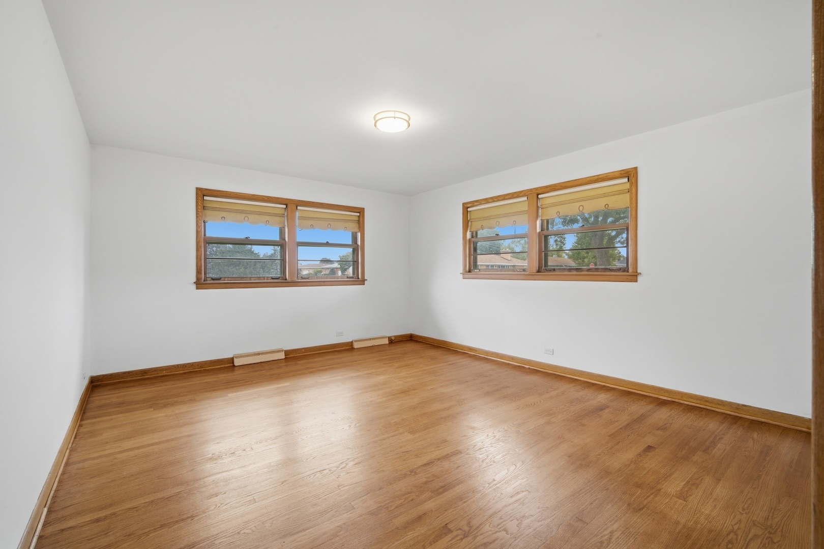 Empty room with wood floors and trim, featuring two sets of windows with raised shades.