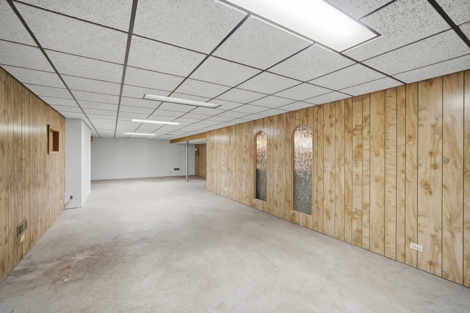 Empty lower-level room with wood-paneled walls, concrete floor, and suspended ceiling.