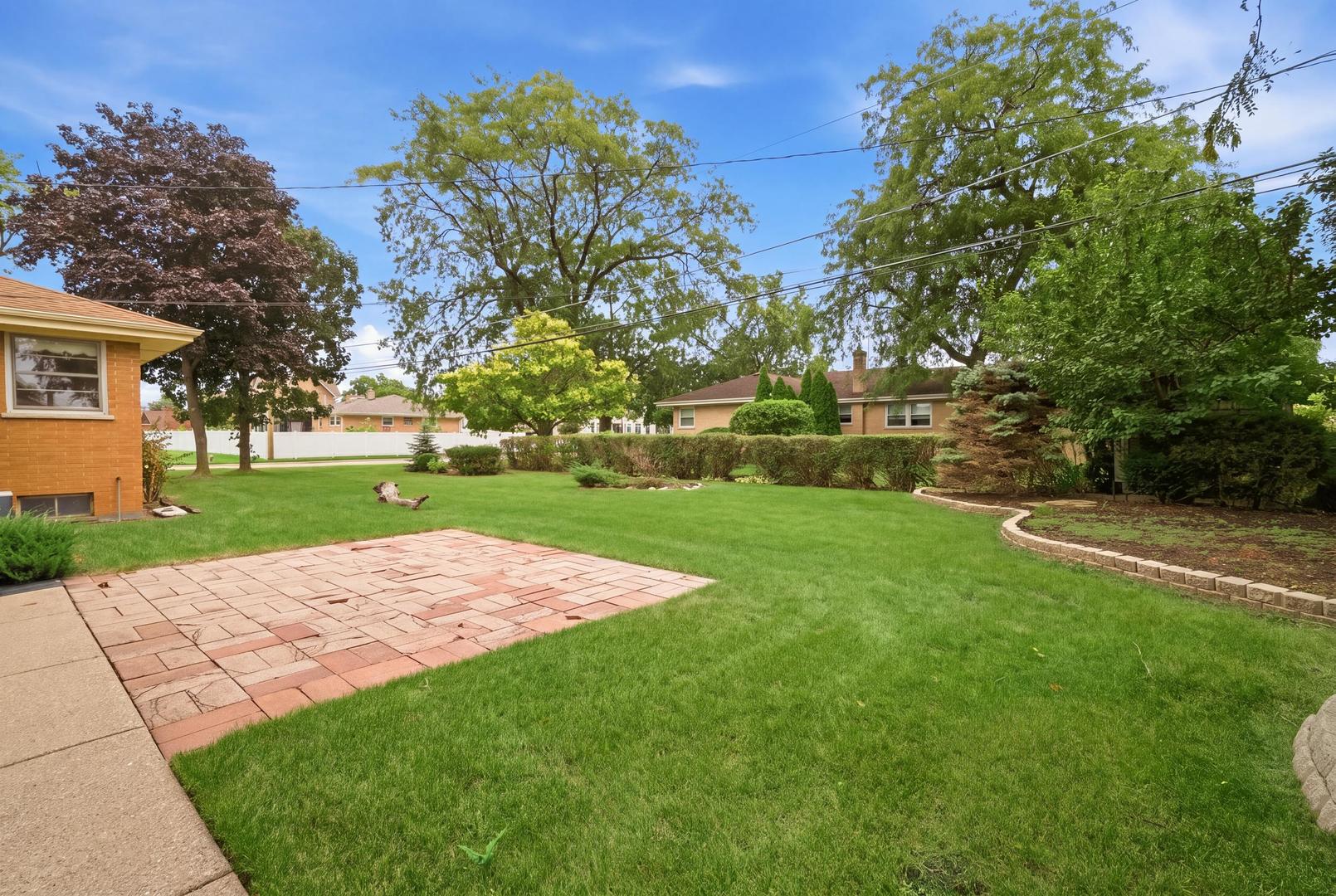 Landscaped backyard with a brick patio, broad lawn, mature trees, and trimmed hedges.