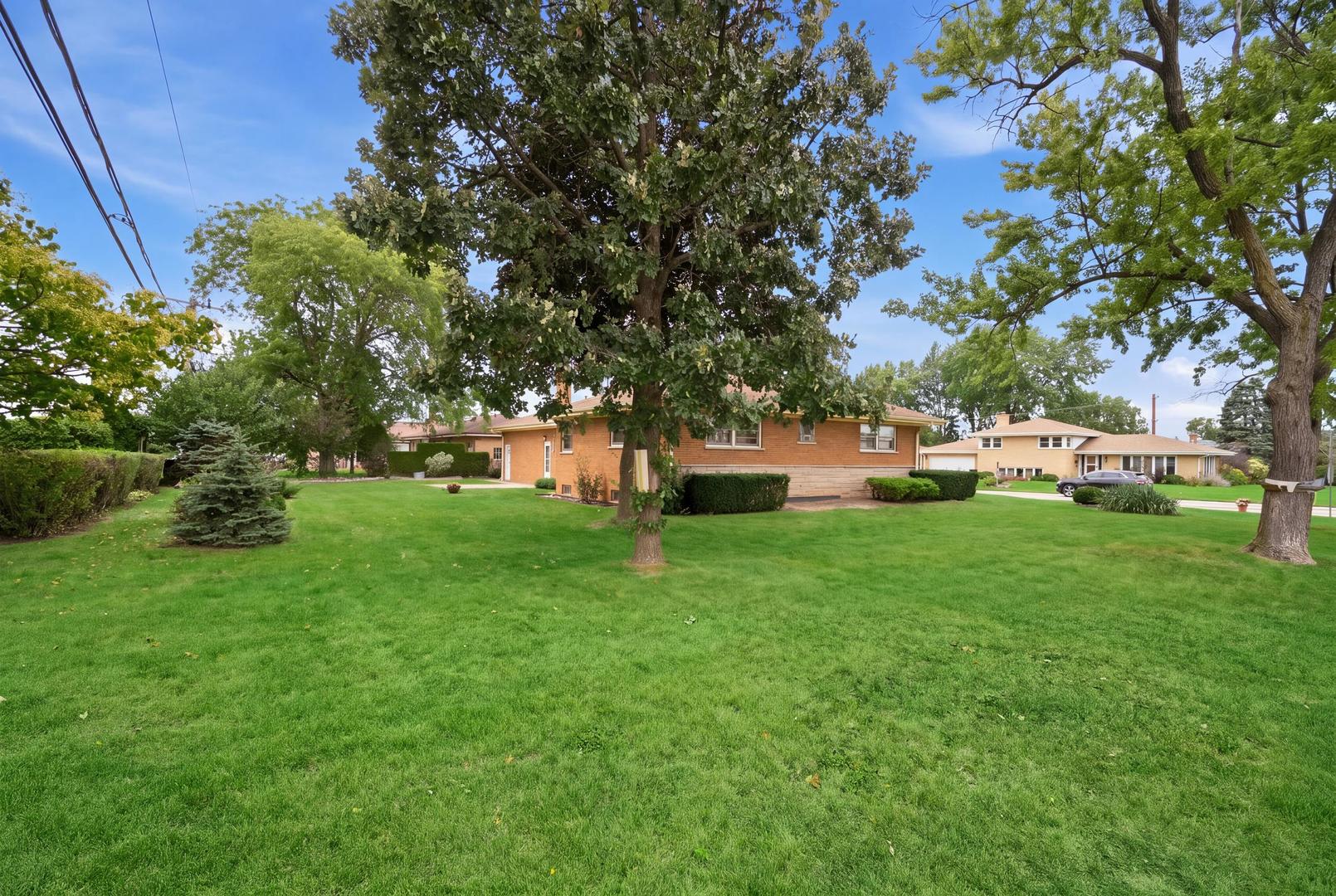 Brick ranch home partly screened by a large shade tree, with a broad green lawn.