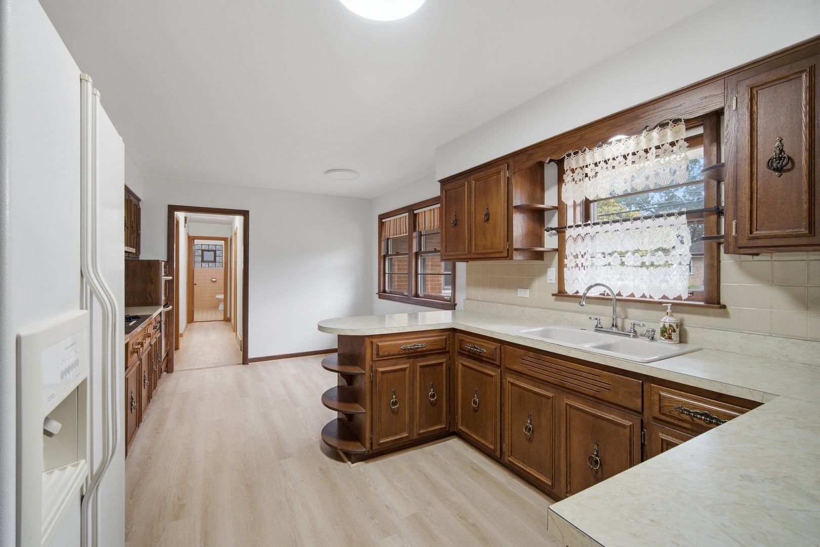 Kitchen with dark wood cabinets, pale countertops, and a double sink beneath lace curtains.