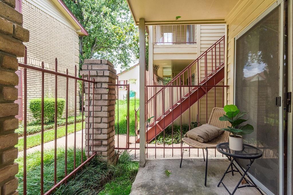 Covered apartment patio with a wicker chair, sliding glass door, red railing, and exterior staircase.