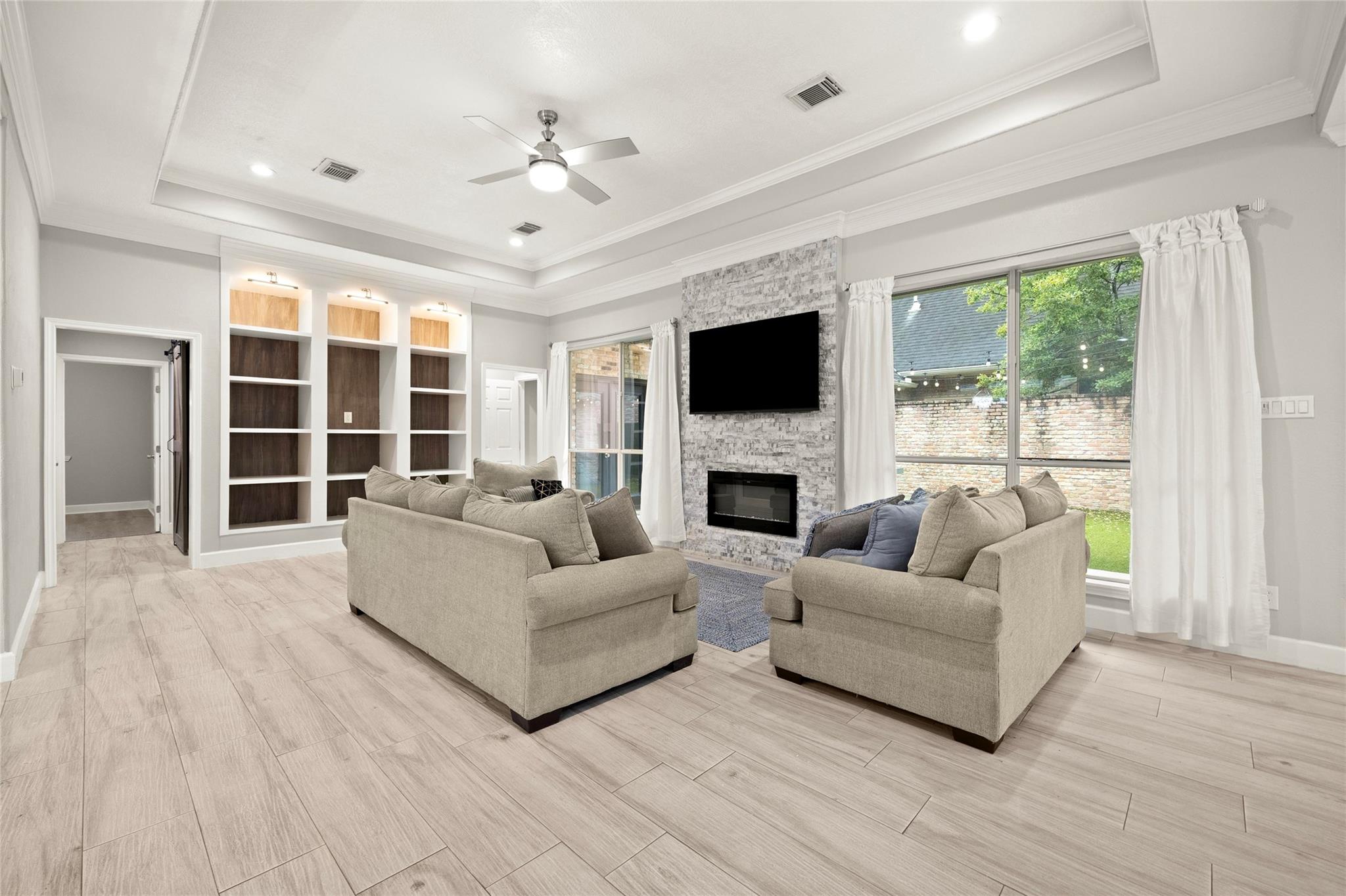 Living room with two beige sofas, a stone fireplace, built-in shelves, and large windows overlooking the yard.
