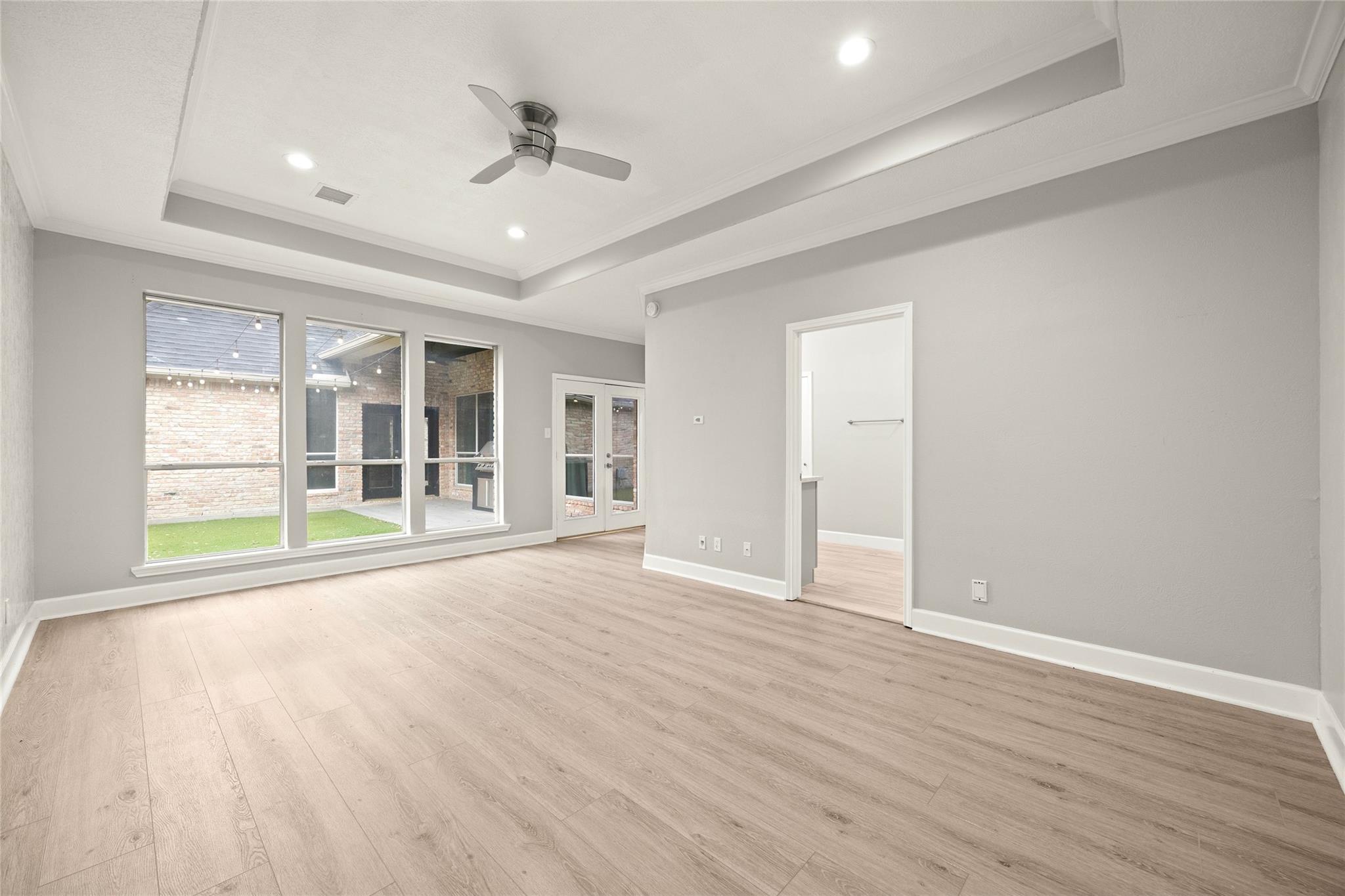 Empty living room with a tray ceiling, ceiling fan, and large windows overlooking a courtyard.
