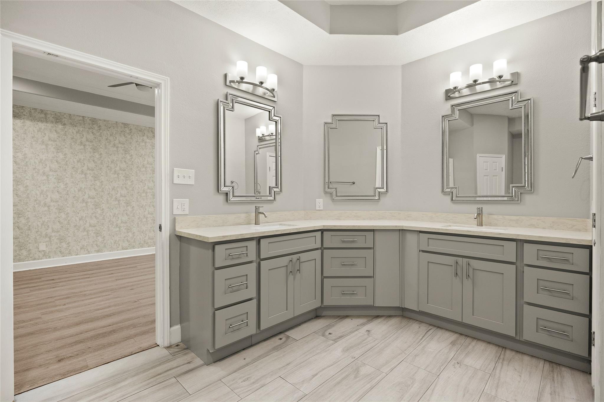 Bathroom with an L-shaped gray double vanity, three framed mirrors, and pale wood-look tile flooring.