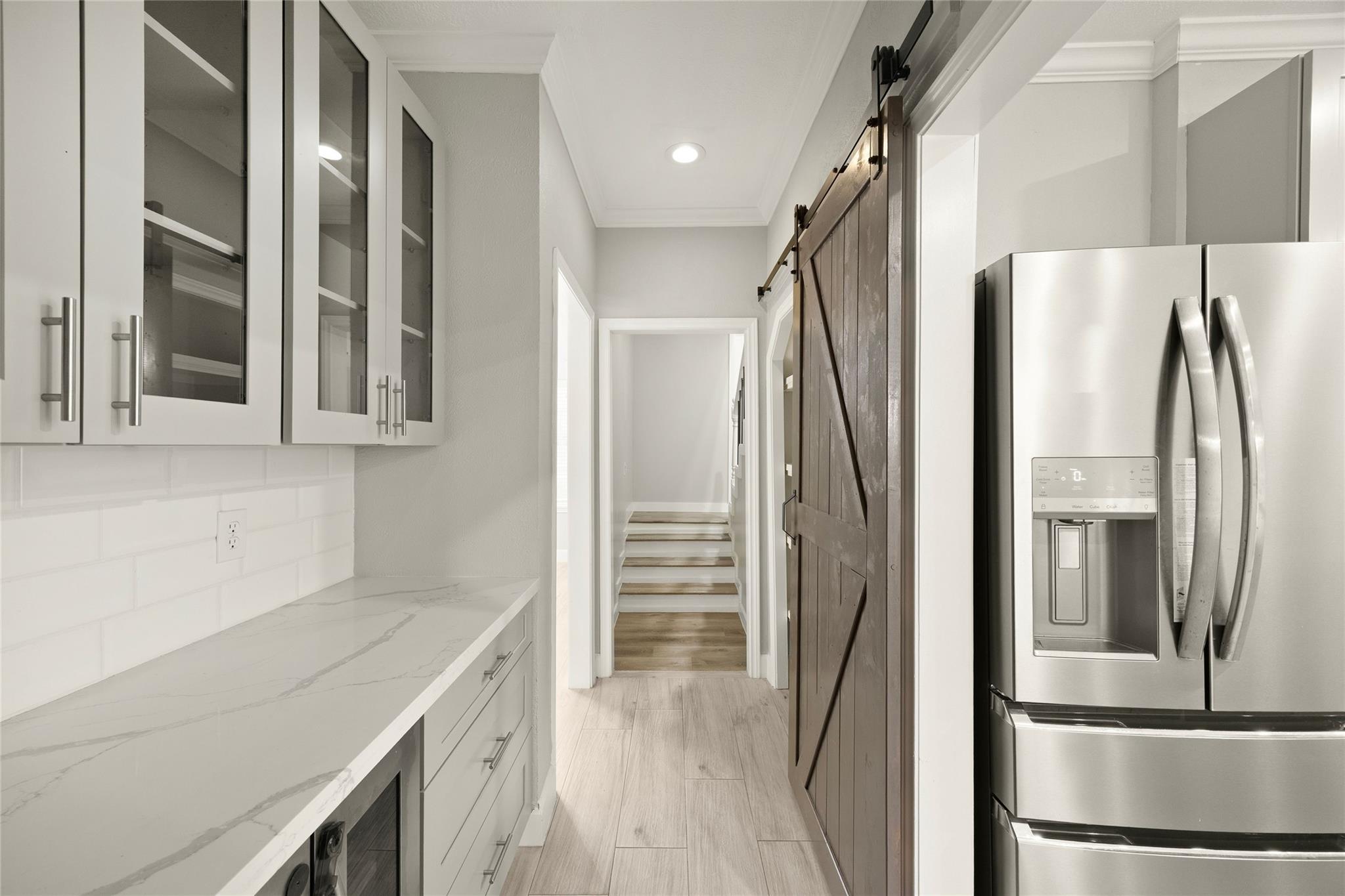 Modern kitchen corridor with white cabinets, a stainless refrigerator, dark barn door, and stairs beyond.