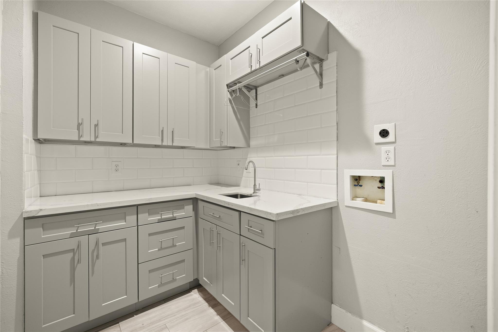 Laundry room with gray cabinets, white tile backsplash, utility sink, and washer hookups.
