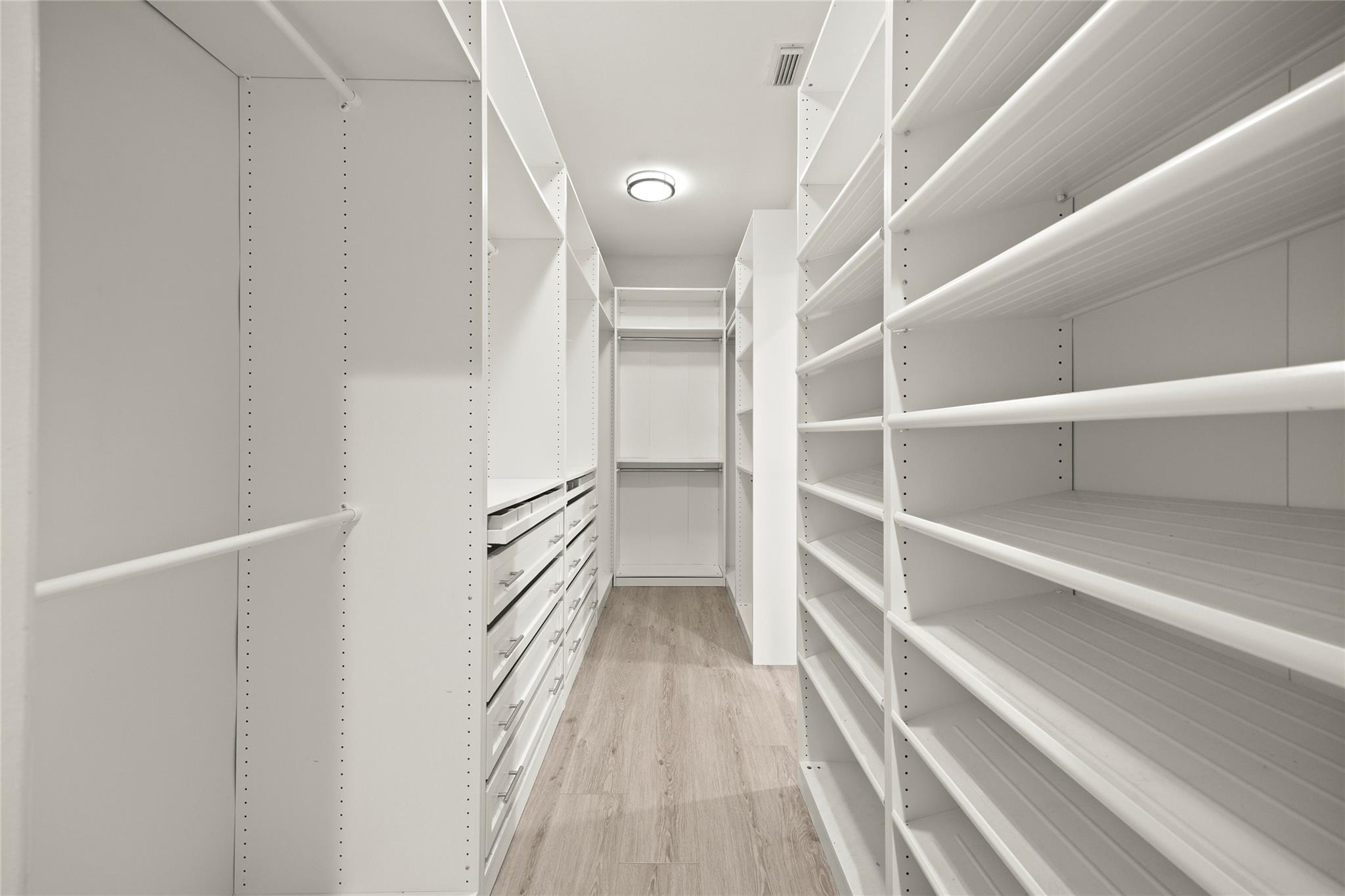 Walk-in closet with white built-in shelving, drawers, and light wood-look flooring.