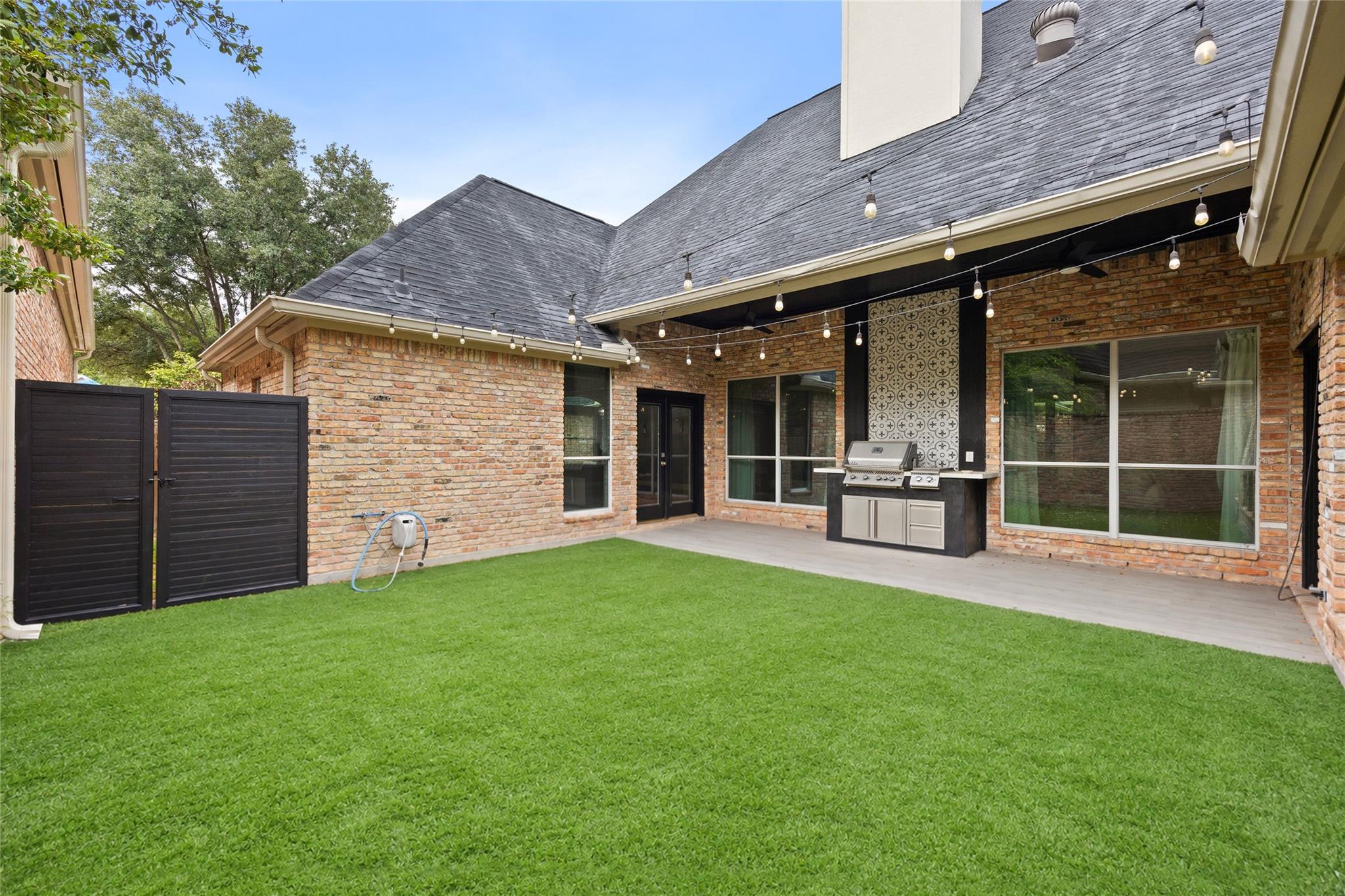 Brick home’s backyard with a green lawn, covered patio, string lights, and built-in grill.