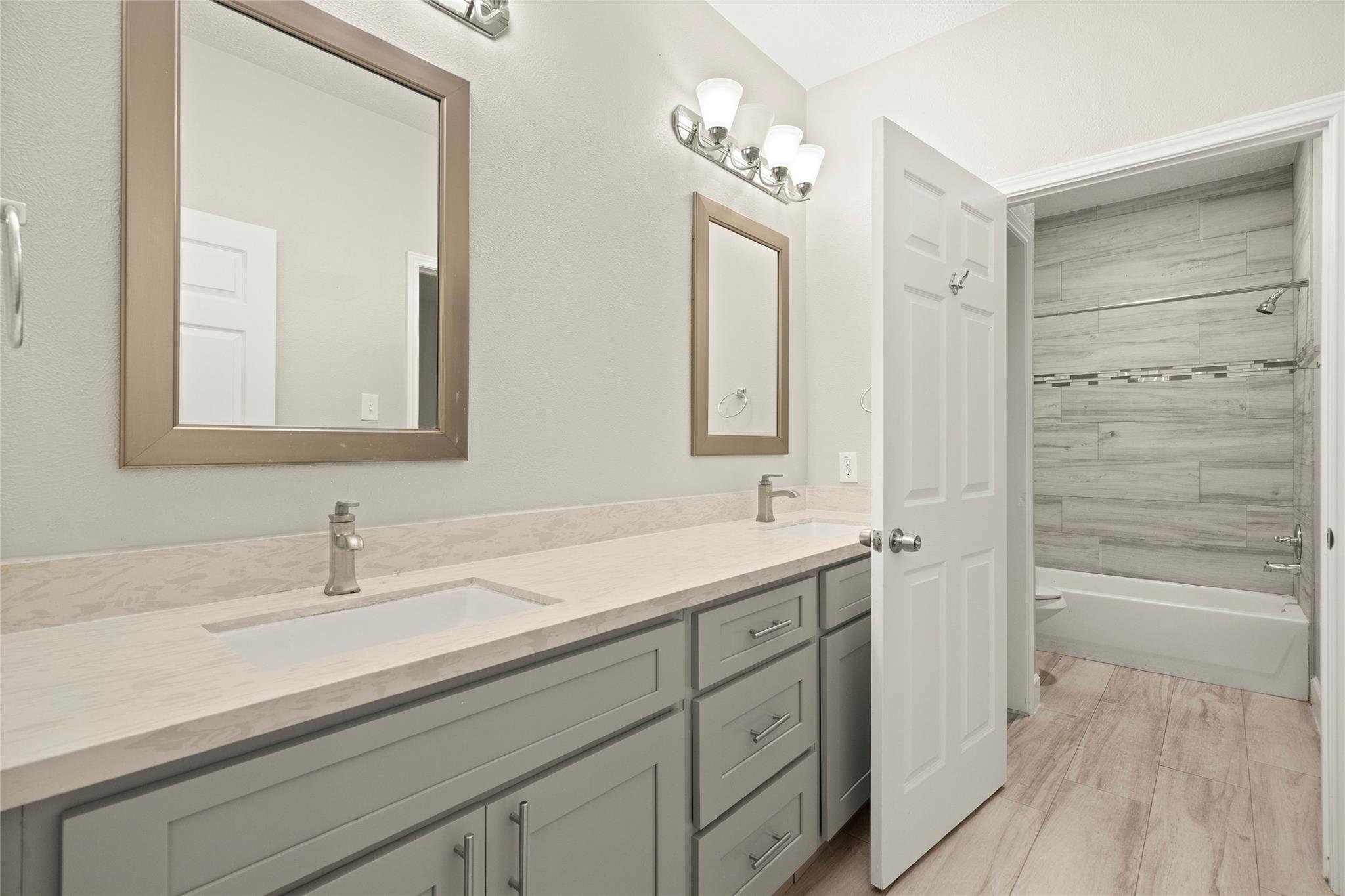 Bathroom with a long double vanity and a tub-shower alcove lined in gray tile.