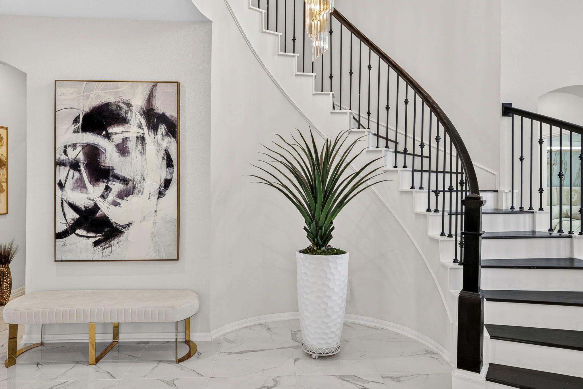 Entryway with a curved black-and-white staircase, potted plant, abstract artwork, and upholstered bench.